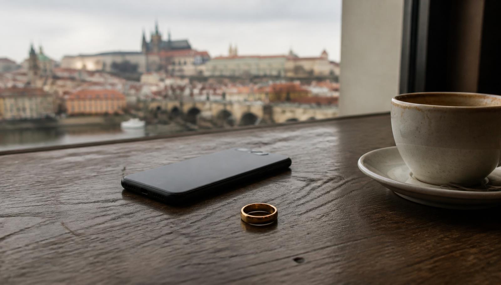 Wedding ring and a face-down smartphone on a café table with a blurred Prague skyline through the window
